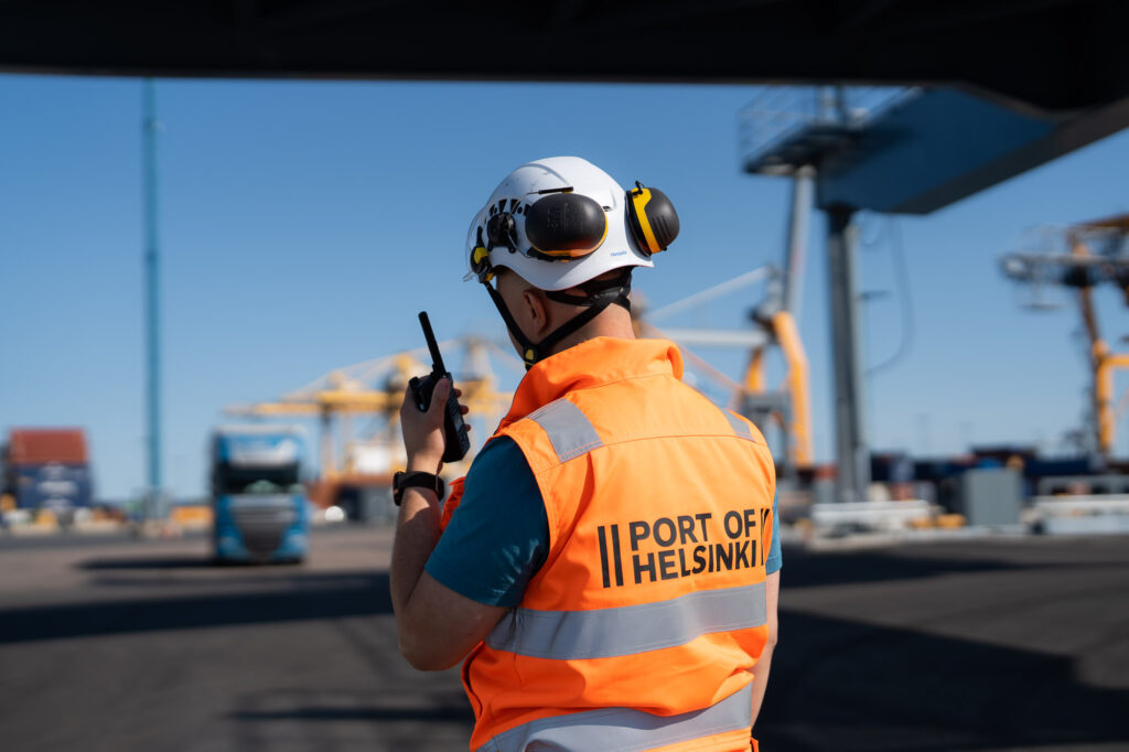 Henkilö puhuu radiopuhelimeen Helsingin Sataman turvaliivi ja kypärä päällä satamassa.