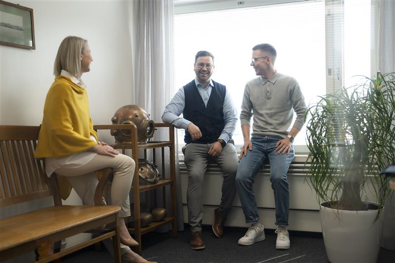 Kaksi ihmistä nojaa ikkunalautaan ja kolmas istuu pöydän reunalla iloisena